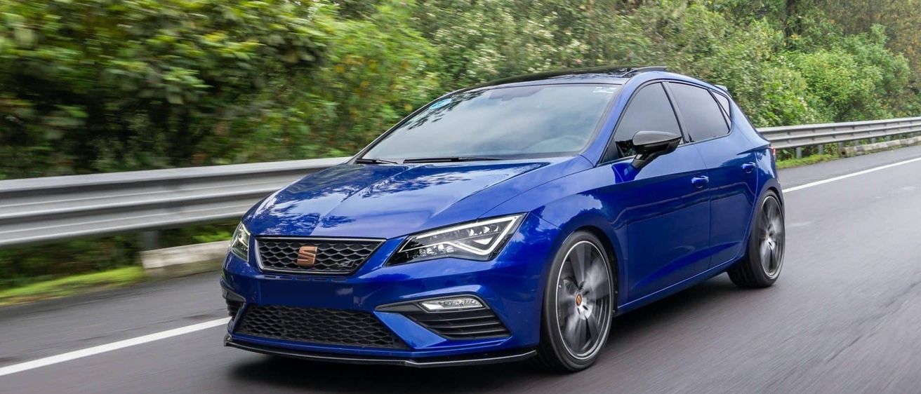 Ein blauer Sportwagen fährt auf einer Straße mit einem Metallschutzgeländer auf der linken Seite. Das Auto ist von Bäumen und Büschen umgeben.