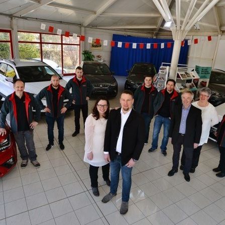 Eine Gruppe von Menschen in Jacken und Brillen steht in einer Ausstellungshalle mit mehreren Autos, darunter rote, weiße und schwarze Fahrzeuge.