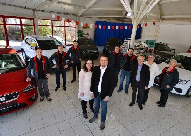 Eine Gruppe von Menschen in Jacken und Brillen steht in einer Ausstellungshalle mit mehreren Autos, darunter rote, weiße und schwarze Fahrzeuge.