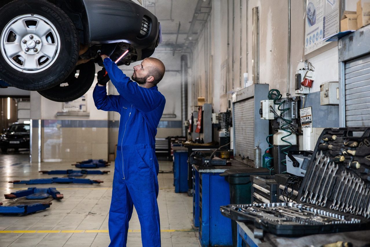 A man in a blue jumpsuit works on a car in a garage, holding a flashlight.
