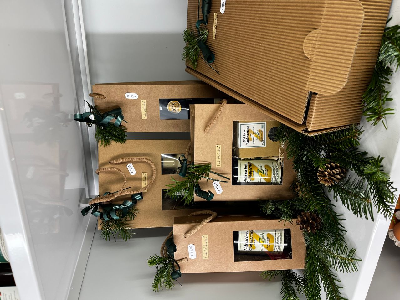 Ein Stapel brauner Kartonboxen mit Bändern und Grünpflanzen auf einer weißen Oberfläche. Die Boxen enthalten Produkte mit Etiketten wie 'Stie richer' und 'Eisberg'.