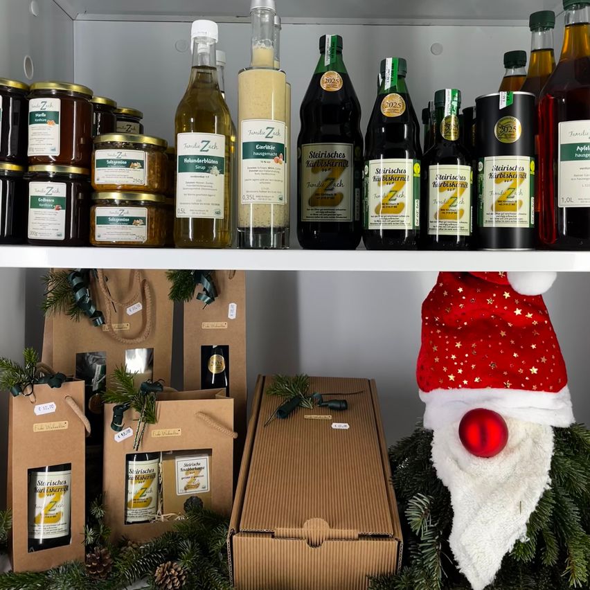 Ein weißer Regalboden zeigt verschiedene Flaschen und Gläser, darunter Produkte mit den Aufschriften 'Stirsches' und 'Zah'. Unter den Flaschen befinden sich verpackte Geschenkboxen und eine dekorative Weihnachtsmütze. Die Szene ist festlich mit Tannenzapfen und Grünpflanzen.