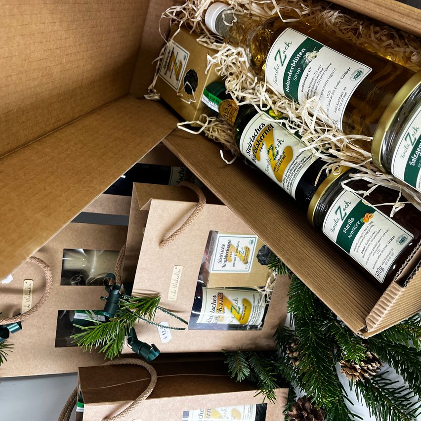 Ein Pappkarton gefüllt mit Flaschen verschiedener Flüssigkeiten, darunter Weißwein, Zitronensaft und Honig. Der Karton ist von Tannennadeln und Tannenzapfen umgeben, möglicherweise für ein weihnachtliches Geschenk.