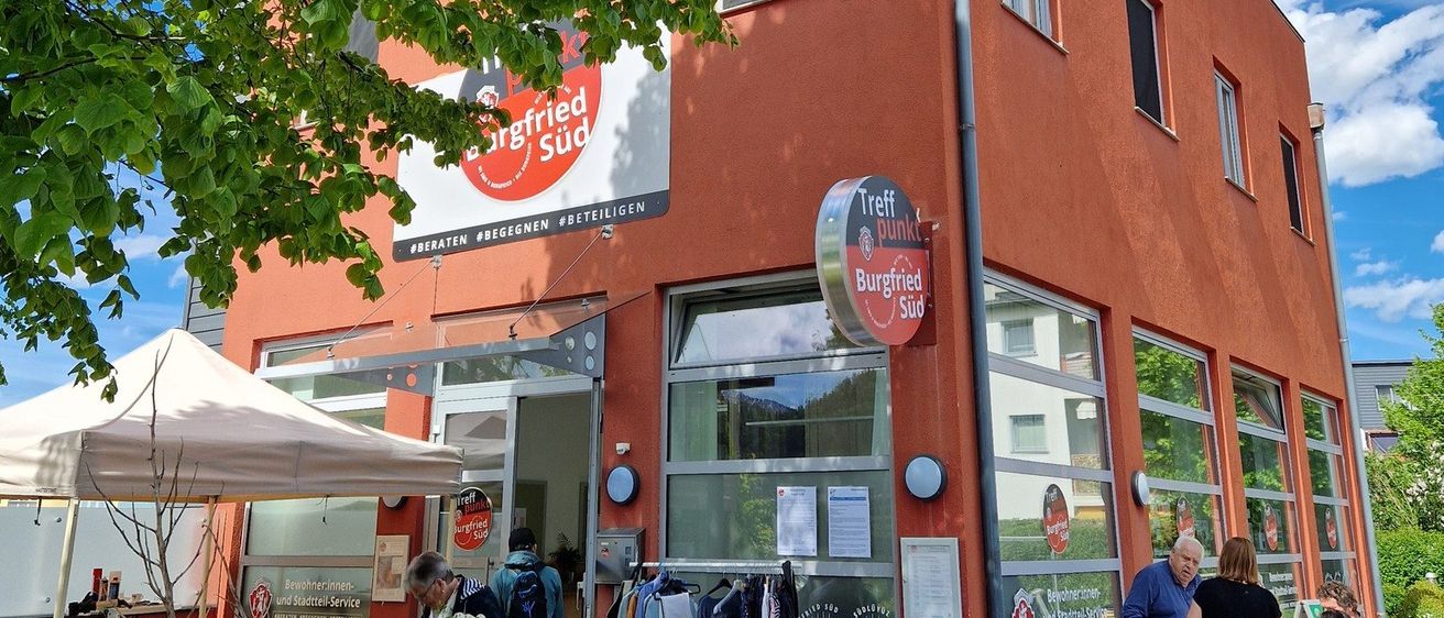 Ein rotes Gebäude mit einem Schild, das 'Urfried Sud' liest, hat eine Gruppe von Menschen draußen. In der Nähe sind Fahrräder geparkt, und es gibt einen Baum mit grünen Blättern darüber.