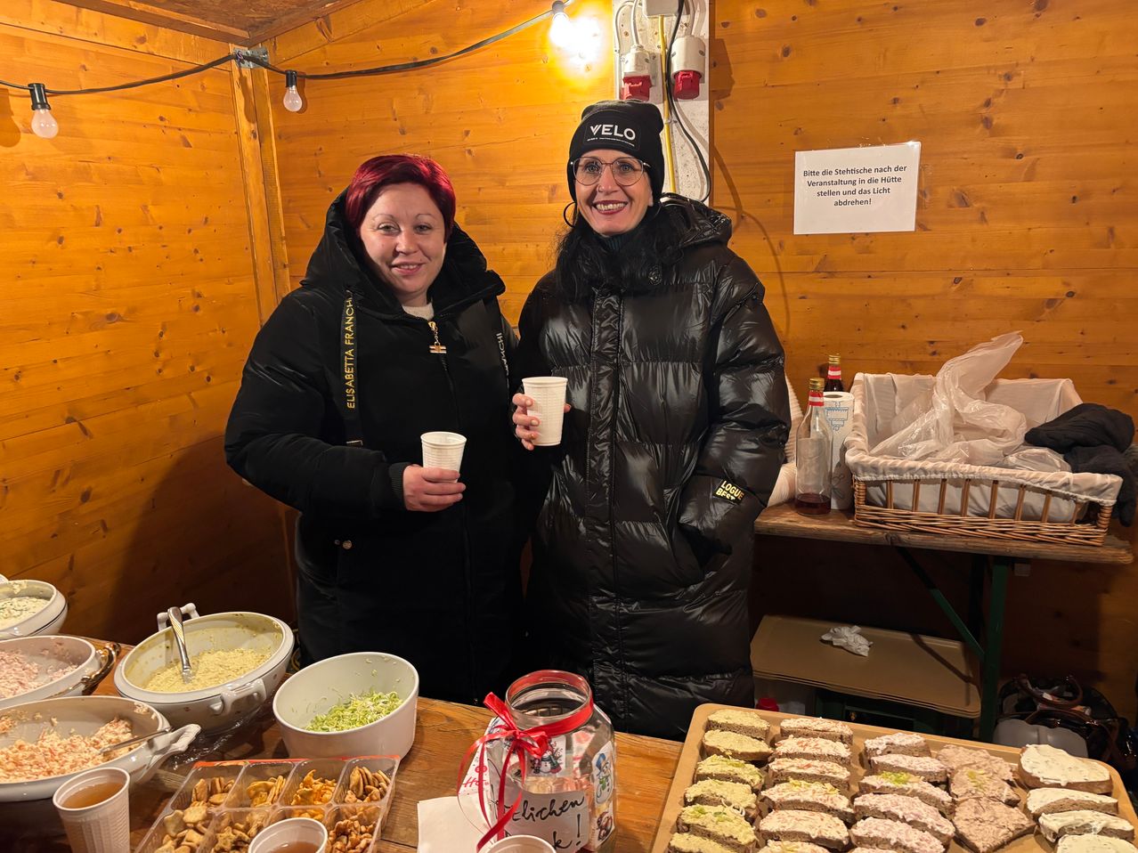 Zwei Frauen stehen nebeneinander, halten Tassen und lächeln. Dahinter eine Holzwand mit einem Schild, ein Tisch mit Essen und einem Korb. Sie tragen schwarze Jacken und Mützen.