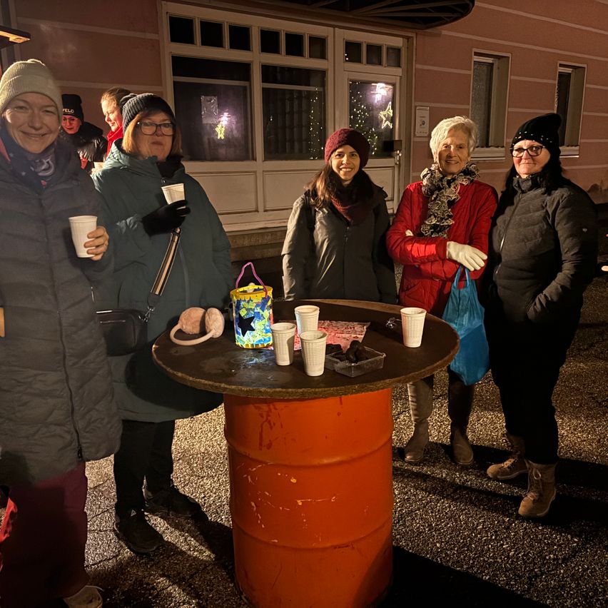Fünf Frauen, die warm gekleidet sind, stehen nachts draußen um einen Tisch herum, lächeln und halten Tassen. Ein brennendes Kerze und ein Korb sind auf dem Tisch.