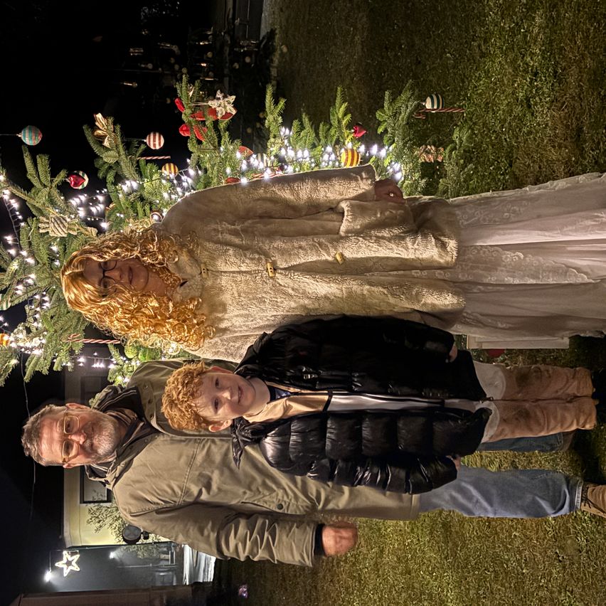 Eine Familie mit drei Personen steht vor einem mit Lichtern, Ornamenten und Grün dekorierten Weihnachtsbaum. Die Frau trägt ein weißes Kleid und der Junge einen schwarzen Mantel. Der Mann trägt einen braunen Mantel. Sie posieren für ein Foto.