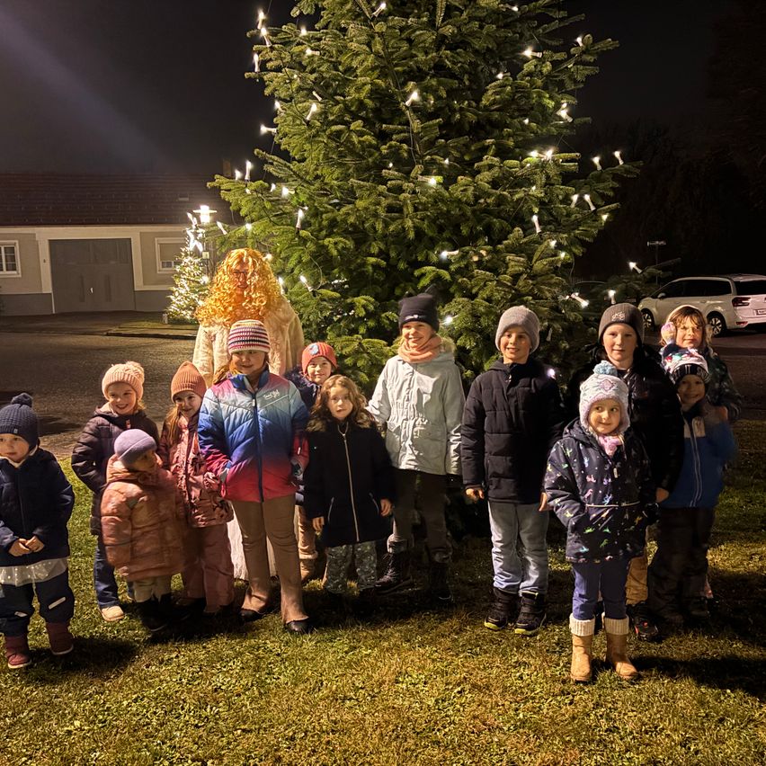 Eine Gruppe von Kindern und zwei Erwachsenen steht nachts vor einem beleuchteten Weihnachtsbaum. Sie sind warm gekleidet.