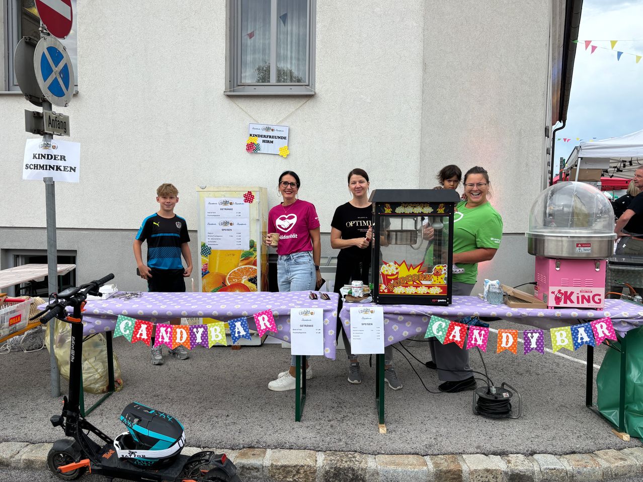 Eine Gruppe von Menschen steht hinter einem Tisch mit einem Candy-Bar-Banner, einer Popcornmaschine und einem Schild. Ein Junge steht links.