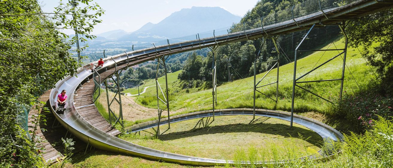 Eine Person auf einer Stahlrutsche kurvt durch die Hügel, umgeben von grünem Gras und Bäumen, mit Bergen in der Ferne.