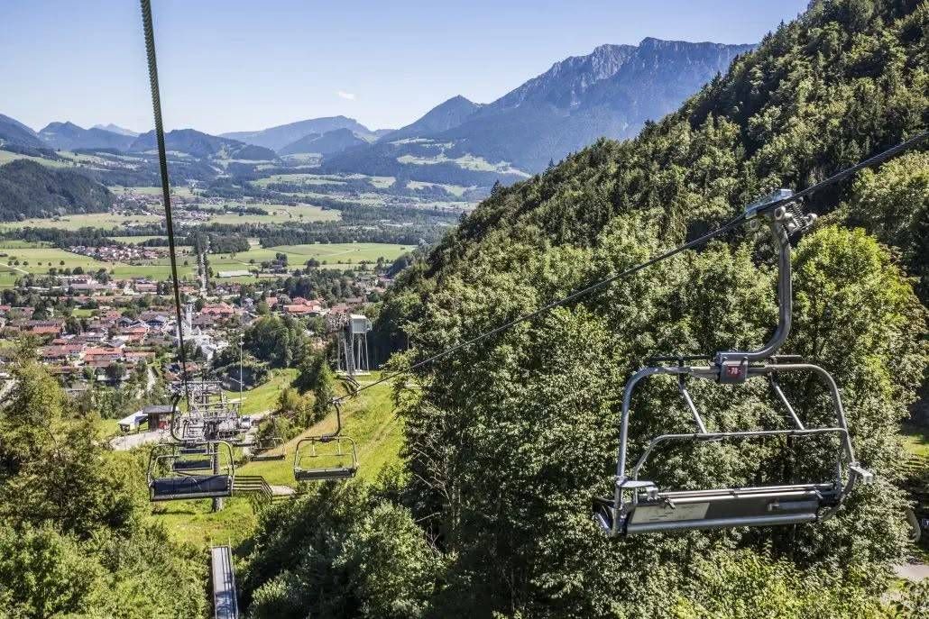 Eine Skiliftbahn erklimmt einen Berg mit Blick auf ein Tal und Häuser unten. Bäume und Berge füllen den Hintergrund.