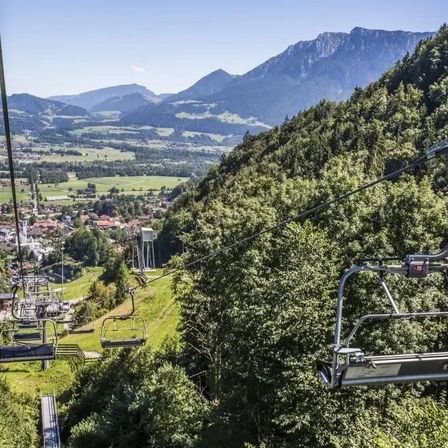 Eine Skiliftbahn erklimmt einen Berg mit Blick auf ein Tal und Häuser unten. Bäume und Berge füllen den Hintergrund.