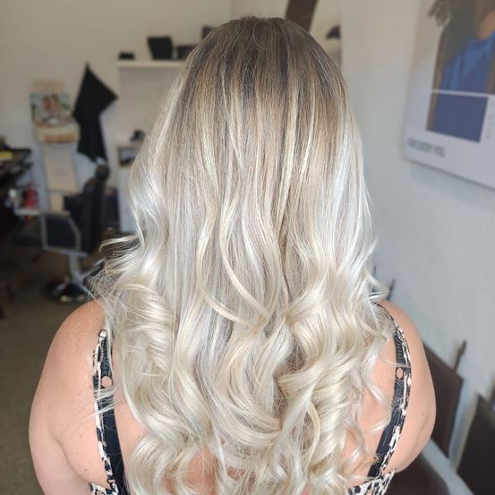 Eine Frau mit langen, welligen, blonden Haaren in einem Salon. Im Hintergrund sieht man eine weiße Wand und ein Poster.