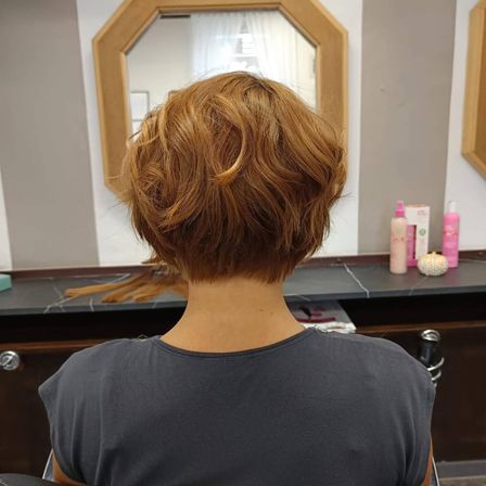 Eine Frau mit kurzen, gelockten roten Haaren sitzt in einem Friseurstuhl. Hinter ihr befindet sich ein Spiegel, der ein Fenster und Haarpflegeprodukte reflektiert.