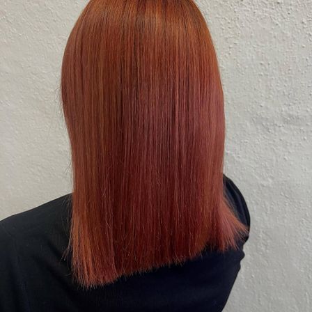 Eine Frau mit langen, glänzenden, roten Haaren, glatt gestylt, Rückansicht, vor einer weißen Wand.
