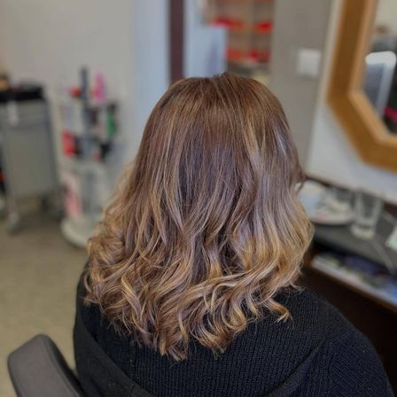 Eine Frau mit blonden, gewellten Haaren sitzt in einem Stuhl in einem Salon. Sie trägt ein schwarzes Oberteil. Hinter ihr spiegelt ein Spiegel einen Schreibtisch.
