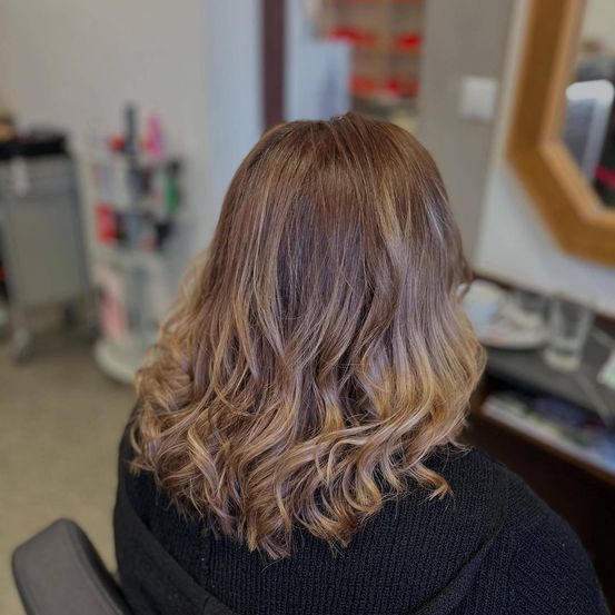Eine Frau mit blonden, gewellten Haaren sitzt in einem Stuhl in einem Salon. Sie trägt ein schwarzes Oberteil. Hinter ihr spiegelt ein Spiegel einen Schreibtisch.