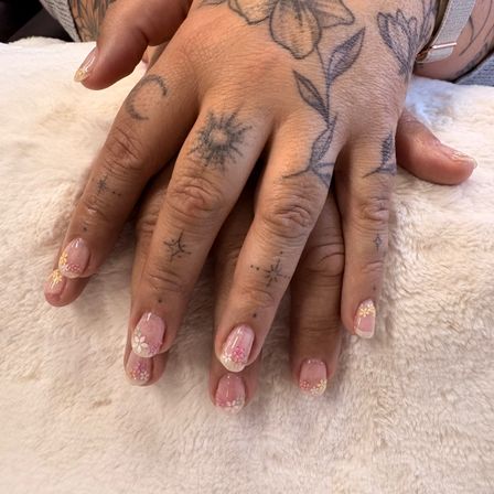 Zwei Hände mit Blumen- und Sternentattoos auf den Knöcheln und mit rosa und weißen Blumendesigns lackierten Nägeln ruhen auf einem weißen Pelztuch.