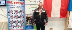 Ein Mann steht vor einem blauen Banner mit der Aufschrift 'Baden bewegt' und einem weißen Banner mit dem Logo von Physioteam Anne Sass. Er trägt eine schwarze Jacke mit einer goldenen Medaille um den Hals. Hinter ihm weht eine rot-weiße Flagge.