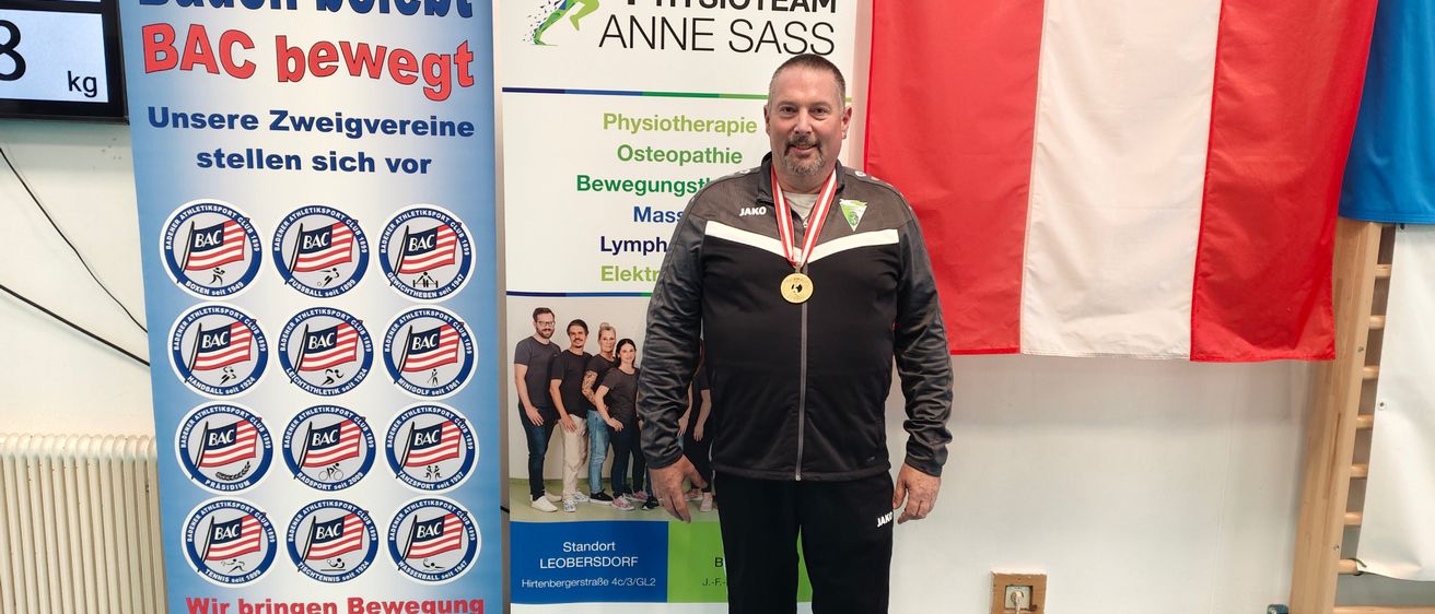 Ein Mann steht vor einem blauen Banner mit der Aufschrift 'Baden bewegt' und einem weißen Banner mit dem Logo von Physioteam Anne Sass. Er trägt eine schwarze Jacke mit einer goldenen Medaille um den Hals. Hinter ihm weht eine rot-weiße Flagge.