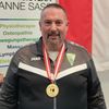 Ein Mann in einem Sportjackett mit einer Goldmedaille posiert vor einem Physioteam Anne Sass-Banner. Dahinter ist eine rot-weiße Flagge.