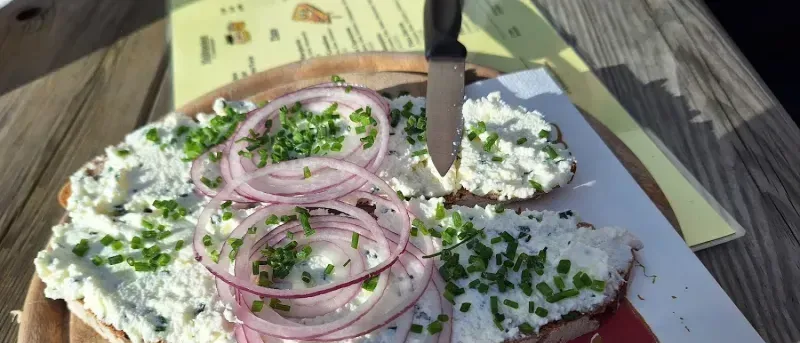 Ein Holzteller mit zwei Brotscheiben, belegt mit Frischkäse, roten Zwiebeln und Schnittlauch. Ein Messer liegt darauf.