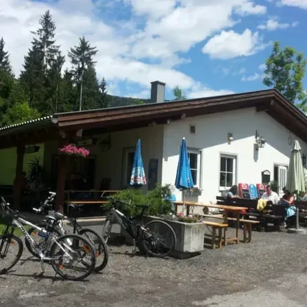 Ein weißes Gebäude mit braunem Dach und einem Terrassenbereich, wo mehrere Fahrräder vor dem Gebäude geparkt sind. Es gibt mehrere Personen, die an Tischen sitzen, und einige Sonnenschirme sind auf den Tischen platziert.
