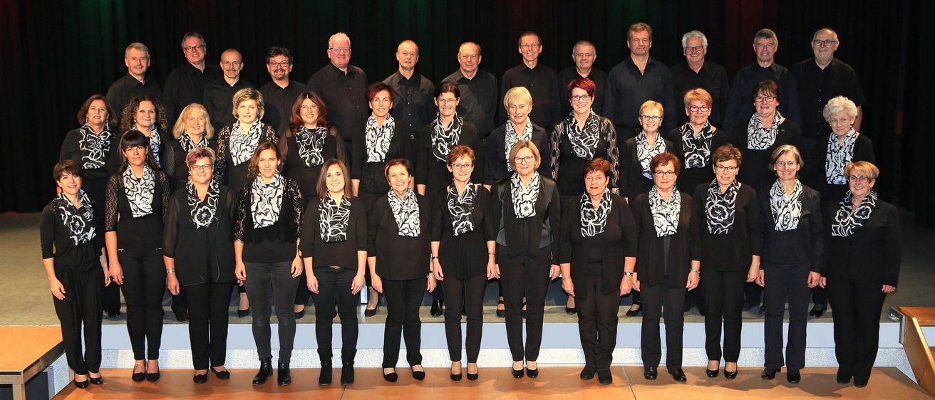 Ein Chor aus Männern und Frauen in Schwarz steht auf der Bühne vor einem grün-roten Vorhang. Sie lächeln und scheinen für ein Foto zu posieren.