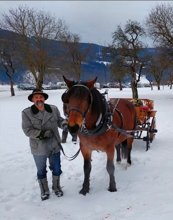 Ein Mann in Winterkleidung steht im Schnee neben einem Pferd, das einen Wagen zieht. Dahinter sind mehrere Autos geparkt. Im Hintergrund befinden sich Bäume und eine entfernte Bergkette.