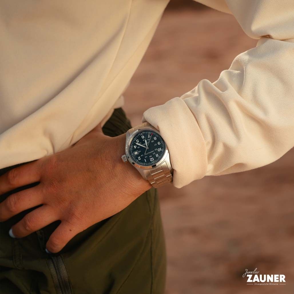 Eine Person trägt eine Armbanduhr am linken Handgelenk, während die rechte Hand in der Hosentasche steckt. Die Uhr hat ein schwarzes Zifferblatt mit silbernen Zeigern und einem silbernen Band.