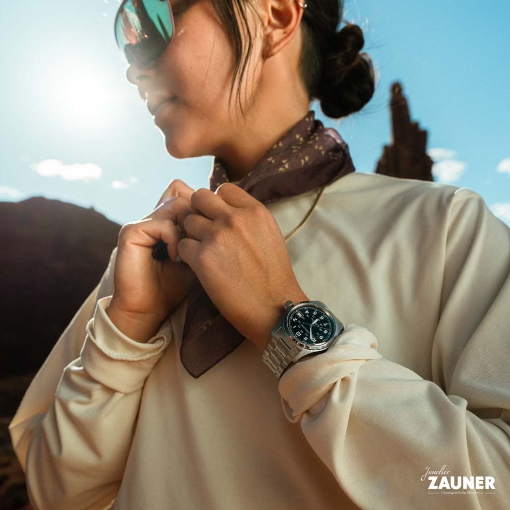 Eine Frau justiert ihren Schal, während sie eine Sonnenbrille und eine Uhr trägt, vor einem Hintergrund von Bergen und klarem Himmel.