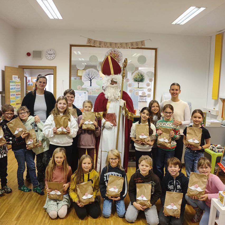 Eine Gruppe von Kindern und Erwachsenen posiert mit dem Heiligen Nikolaus, Geschenktüten in der Hand. Der Raum ist mit einer Geburtstagsdekoration und einem Baum geschmückt.