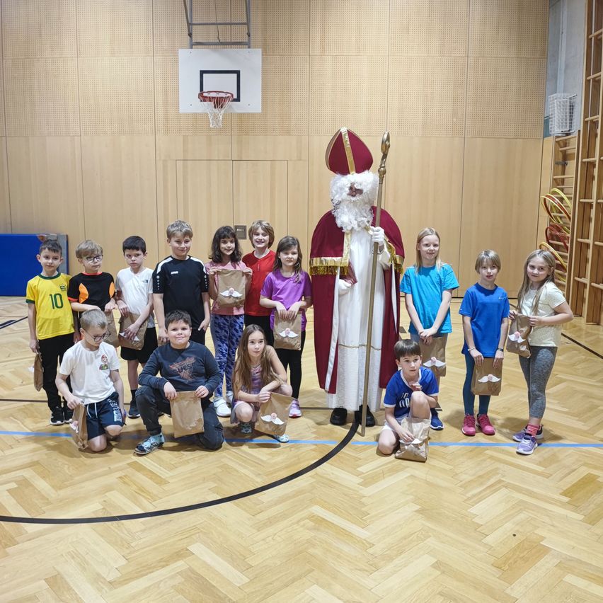 Eine Gruppe von Kindern, in verschiedenen farbigen T-Shirts gekleidet, posiert für ein Foto mit einem Mann in Santa-Claus-Kostüm in einer Turnhalle.