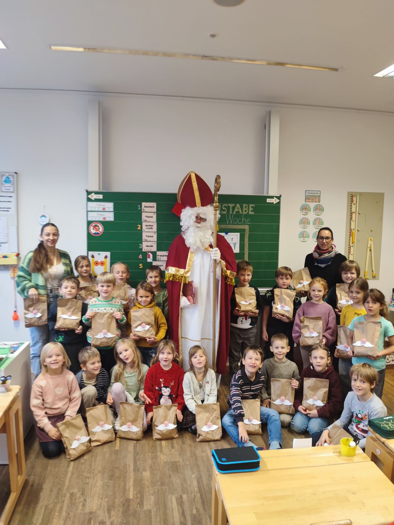 Eine Gruppe von Kindern und Erwachsenen in einem Klassenzimmer, die Papiertüten halten, mit einer als Weihnachtsmann verkleideten Person in der Mitte. Ein Tafel steht hinter ihnen, und eine Frau steht links.