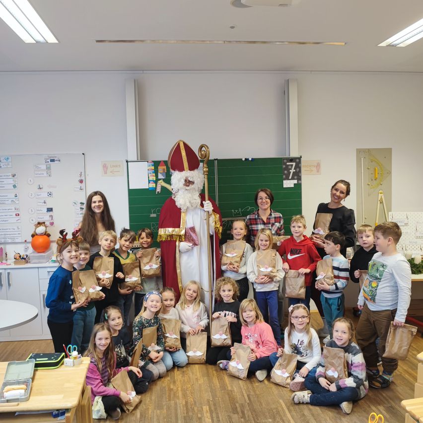 In einem Klassenzimmer posieren Kinder und Erwachsene für ein Foto mit einer als Weihnachtsmann verkleideten Person. Alle Kinder halten Papiertüten, und einige von ihnen tragen Brillen. Das Klassenzimmer hat weiße Wände und Holzböden.
