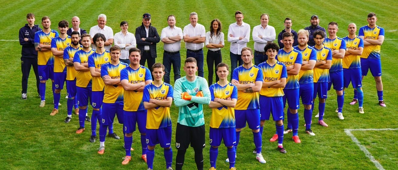Eine Fußballmannschaft in blauen und gelben Trikots posiert für ein Foto auf einem grünen Feld, wobei der Torwart vorne steht. Mitarbeiter in weißen Hemden stehen hinter ihnen.