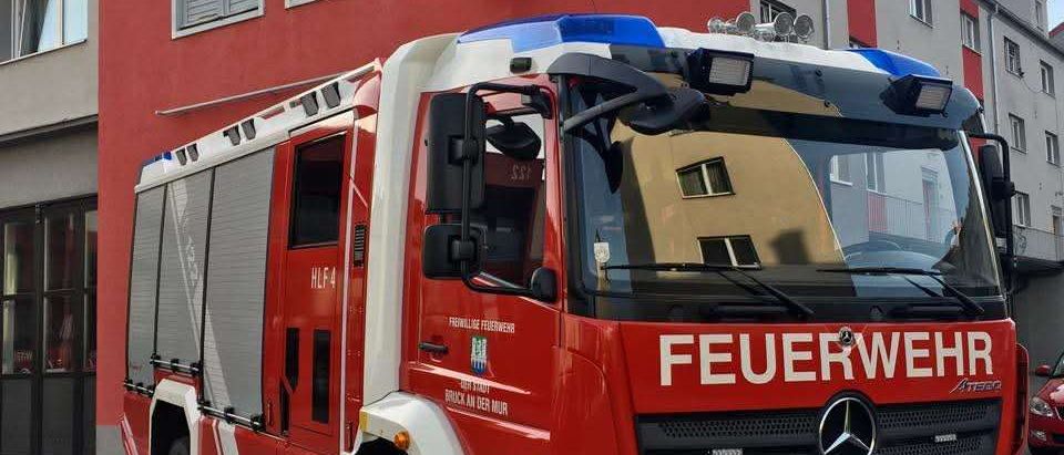 Ein roter und weißer Feuerwehrwagen steht vor einem roten Gebäude mit einer Statue. Der Wagen trägt die Aufschrift 'FEUERWEHR' auf der Frontseite.