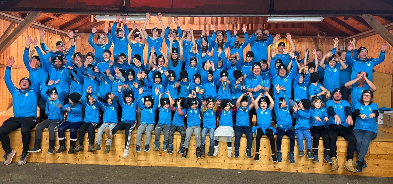 Eine Gruppe von Kindern und Erwachsenen in blauen Hemden und Mützen, die auf einer Holzbank sitzen und stehen, lächeln und ihre Hände heben.