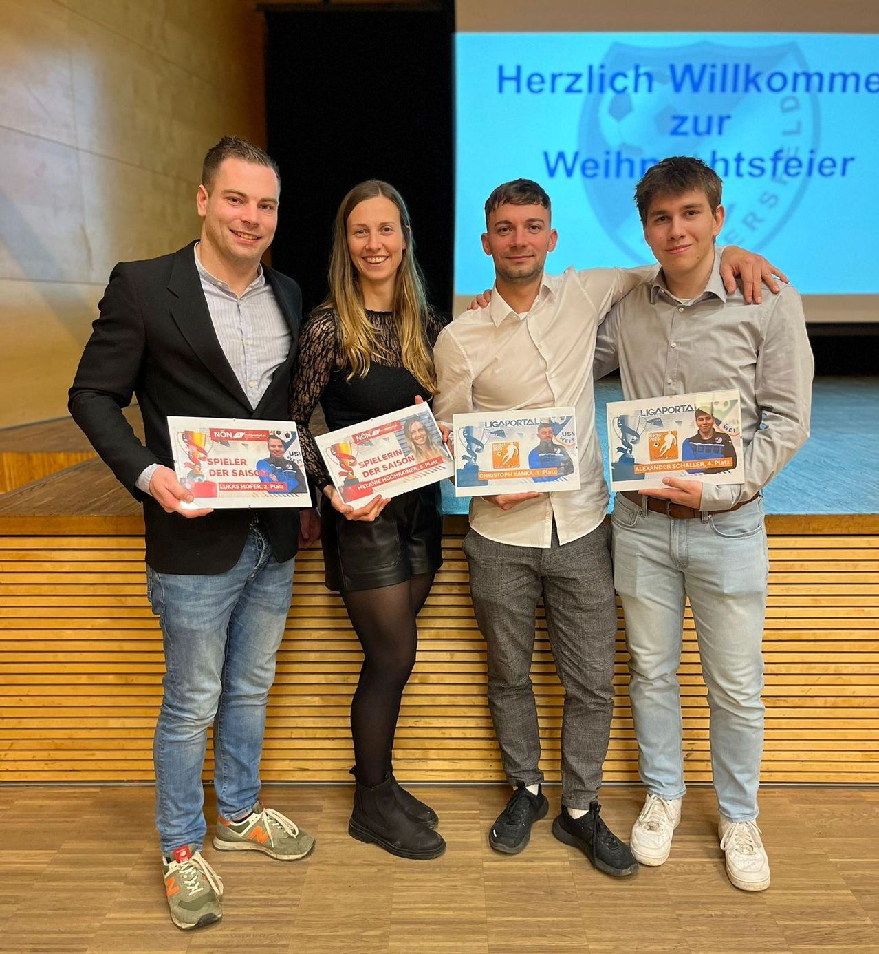 Vier Personen stehen gemeinsam vor einem Podium. Sie halten Urkunden in den Händen. Eine Leinwand hinter ihnen zeigt eine Willkommensbotschaft. Der Mann auf der linken Seite trägt einen schwarzen Anzug, während die anderen lässige Kleidung tragen. Sie scheinen eine Veranstaltung zu feiern.