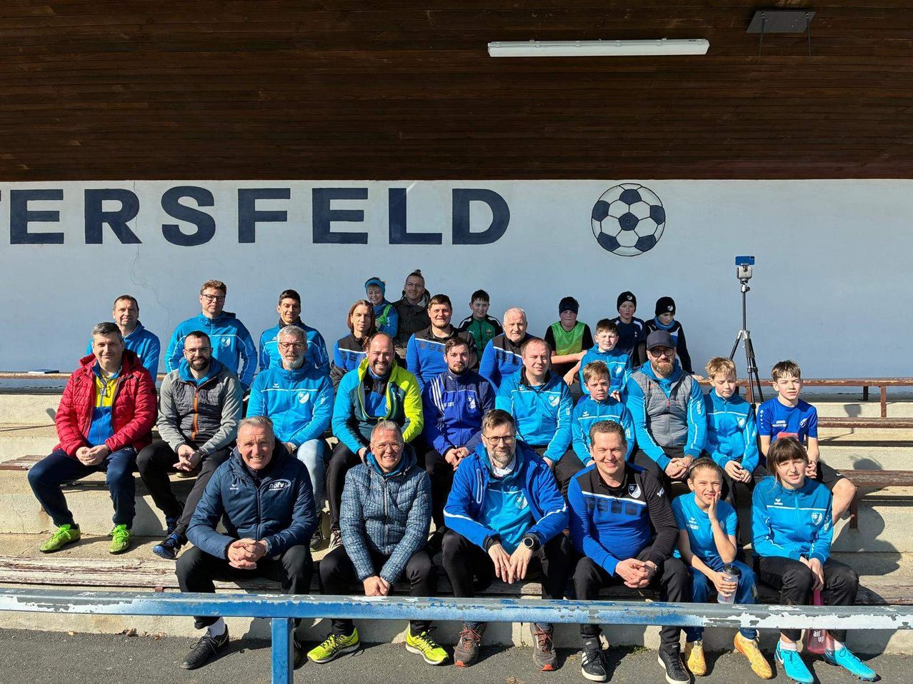Eine Gruppe von Fußballspielern und Trainern, die blaue Uniformen tragen, posiert für ein Foto vor einer Wand mit dem Wort 'Rssfeld' und einem Fußball.