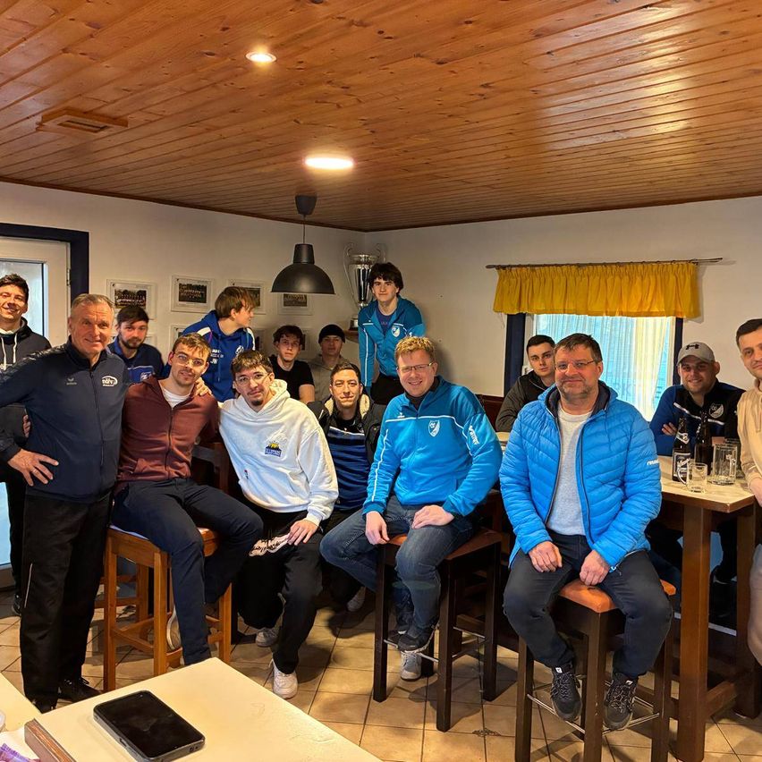 Eine Gruppe von Männern, die in verschiedenen Farben gekleidete Jacken und Hosen tragen, sitzen um einen Tisch herum und posieren für ein Foto in einem Raum mit Holzdecke und Wänden.