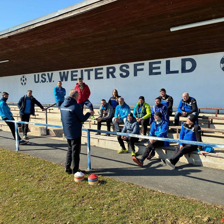 Eine Gruppe von Personen in blauen und gelben Uniformen sitzt auf den Tribünen einer Sportanlage, U.S.V. Weitersfeld. Ein Mann steht in der Nähe des Zauns und hält einen Ball.