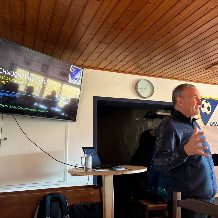 Ein erwachsener Mann spricht an einem Tisch mit Laptop und einem Glas Wasser. Ein großer Monitor ist an der Wand montiert und zeigt Text auf Deutsch. Der Raum hat Holzwände und eine Holzdecke.