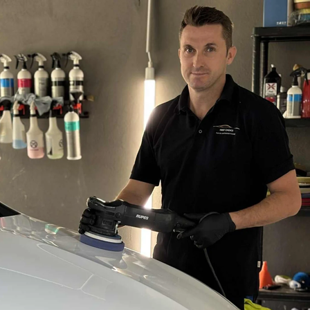 Ein Mann poliert die Motorhaube eines weißen Mercedes-Autos in einer Garage. Er benutzt eine Maschine und trägt Handschuhe. Das Auto hat eine schwarze Kühlergrill und ein Mercedes-Logo.