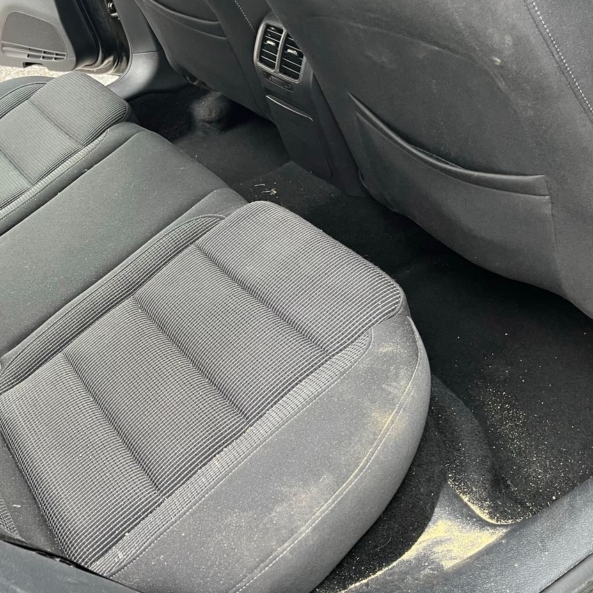 Der Innenraum eines Autos zeigt graue Ledersitze mit sichtbarer Abnutzung, Staub auf dem Boden und eine offene Tür mit einer Lüftungsöffnung.