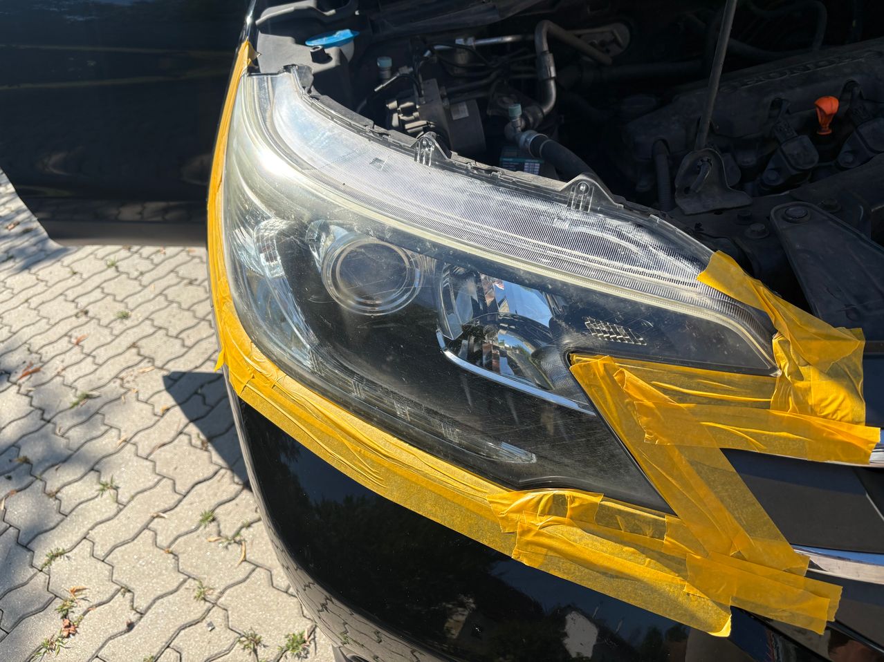 A close-up of a car's headlight with yellow tape covering the edges, the hood is open showing the engine.