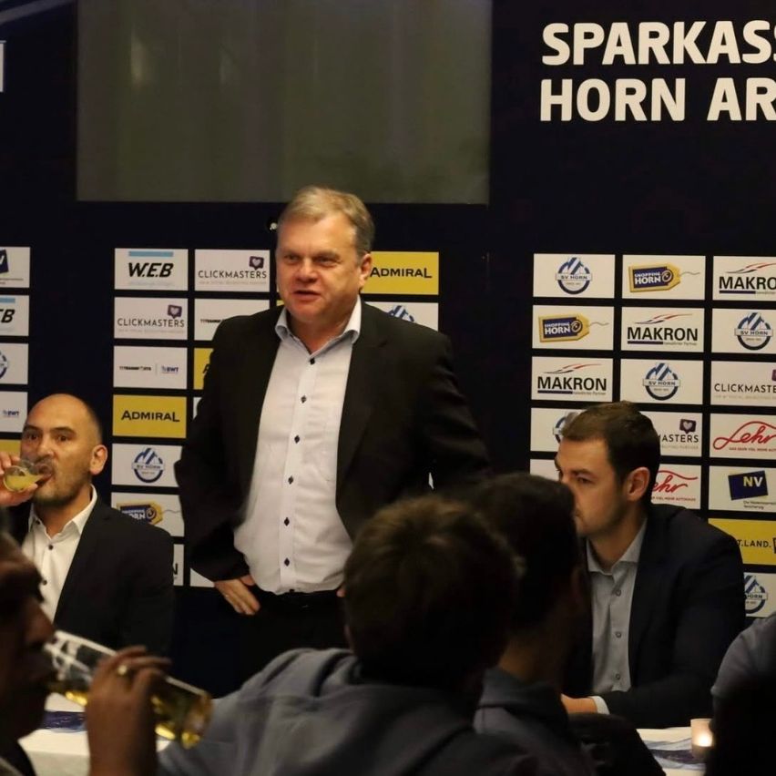 Ein Mann steht vor einer Wand mit verschiedenen Logos und Texten, darunter 'SPARKASSE HORN AR'. Mehrere Menschen sitzen vor ihm und trinken und lauschen.