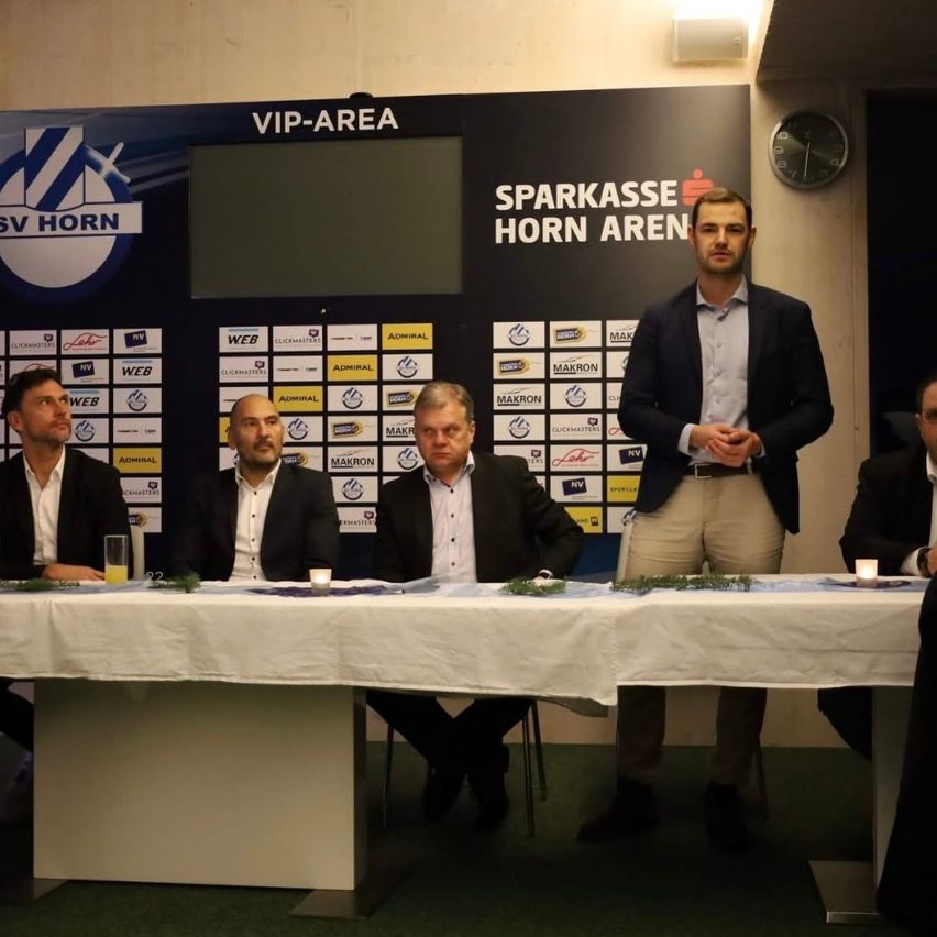 Fünf Männer in Anzügen sitzen an einem langen Tisch mit Kerzen, einer steht auf einem Podium in einem VIP-Bereich. Ein großes Schild hinter ihnen steht Sparkasse Horn Arena.