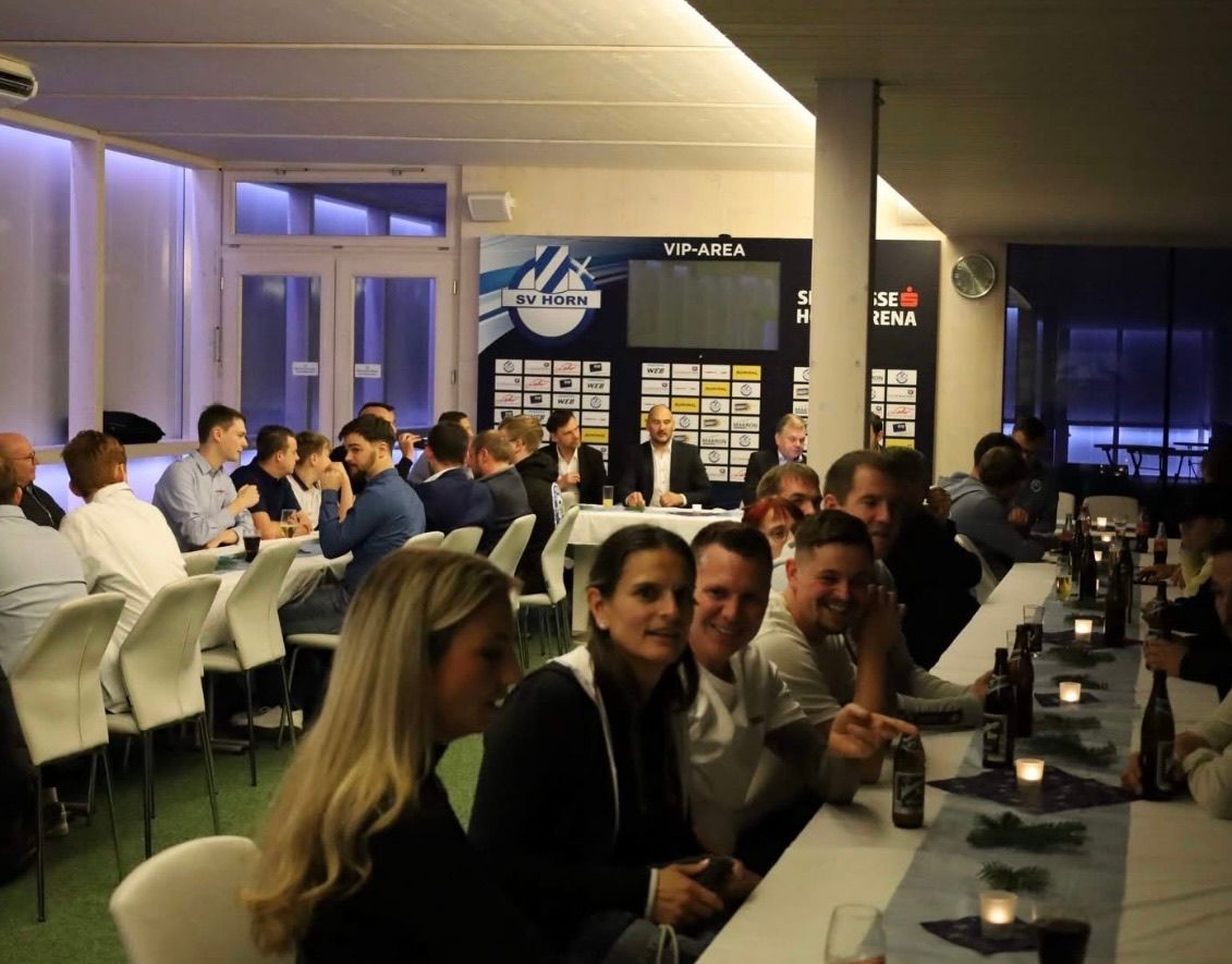 Eine Gruppe von Menschen versammelt sich in einem Restaurant und sitzt um Tische herum. Der Raum hat einen VIP-Bereich mit einem großen Bildschirm und Bannern. Der Raum ist gut beleuchtet mit Lampen, und es gibt Flaschen und Gläser auf dem Tisch.