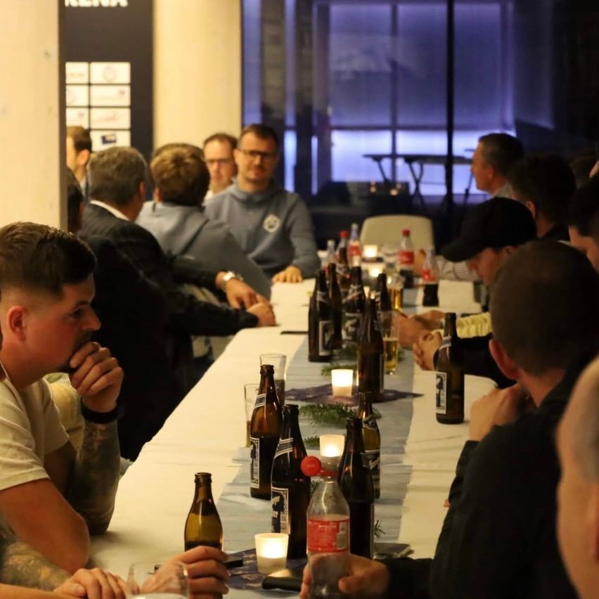 Eine Gruppe von Menschen sitzt um einen langen Tisch mit Flaschen und Gläsern, führt Gespräche, einige halten Getränke. Kerzen und ein Hintergrund mit Text sind sichtbar.